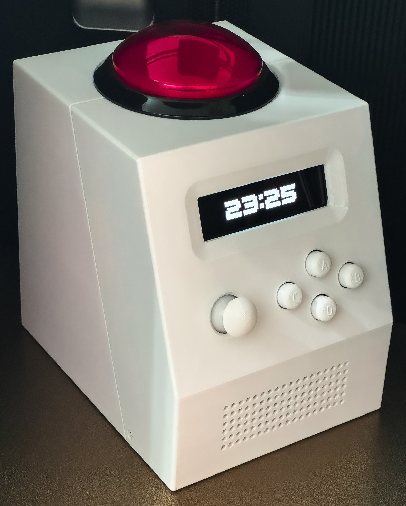 CinderClock on a desk: a white cube with a large red illuminated dome button on top, a wide monochrome display reading 23:25, and a joystick with four round buttons on the raked front face